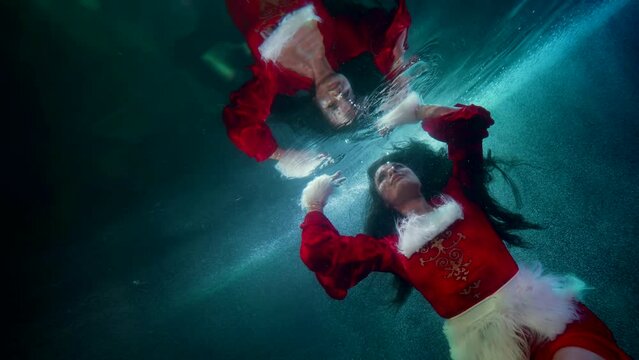 a woman with dark long hair and wearing a red suit with white fur floats in the blue water and touches the surface with hands. the middle plan. the camera is tilted