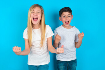 Portrait of two kids boy and girl standing over blue studio background looks with excitement at camera, keeps hands raised over head, notices something unexpected reacts on sudden news.