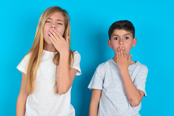 Sleepy two kids boy and girl standing over blue studio background yawning with messy hair, feeling...