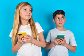 Happy two kids boy and girl standing over blue studio background listening to music with earphones using mobile phone.