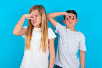 Displeased two kids boy and girl standing over blue studio background plugs nose as smells...