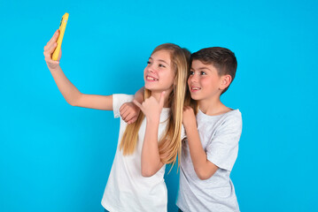 Portrait of a two kids boy and girl standing over blue studio background  taking a selfie to send it to friends and followers or post it on his social media.