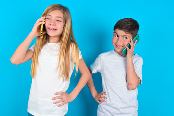 Portrait of successful joyful two kids boy and girl standing over blue studio background talking on mobile phone with friend. Lifestyle and communication concept