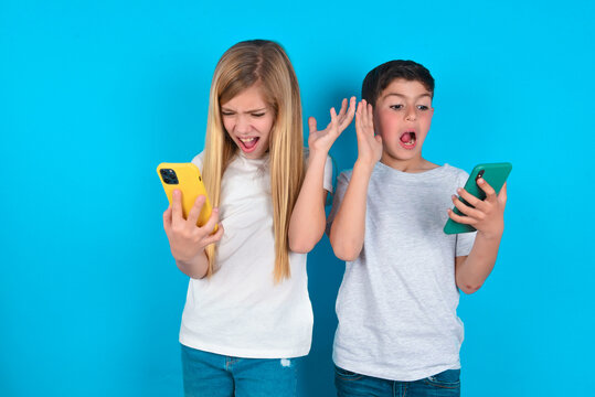 Photo of outraged annoyed two kids boy and girl standing over blue studio background holds cell phone, makes call, argues with colleague,  expresses negative emotions. People and anger.