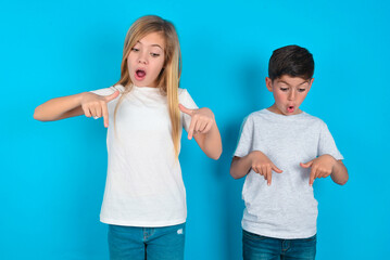 Amazed two kids boy and girl standing over blue studio background points down with fore fingers, opens mouth being shocked. Advertisement concept. - Powered by Adobe
