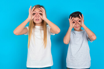 Playful excited two kids boy and girl standing over blue studio background showing Ok sign with both hands on eyes, pretending to wear spectacles.