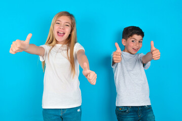 two kids boy and girl standing over blue studio background making positive gesture with thumbs up smiling and happy for success. Looking at the camera, winner gesture. - obrazy, fototapety, plakaty