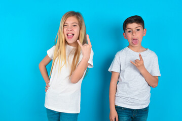 Portrait of a crazy two kids boy and girl standing over blue studio background showing tongue horns up gesture, expressing excitement of being on concert of band.