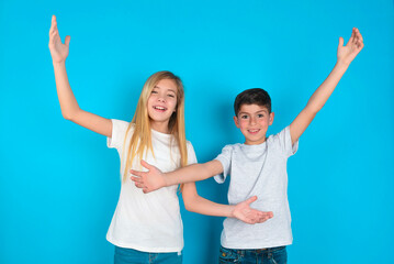 Cheerful two kids boy and girl standing over blue studio background making a welcome gesture raising arms over head. - Powered by Adobe