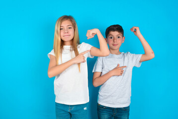 Smiling two kids boy and girl standing over blue studio background raises hand to show muscles,...