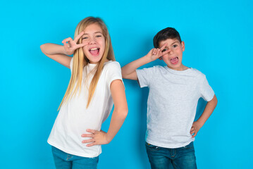 two kids boy and girl standing over blue studio background making v-sign near eyes. Leisure, coquettish, celebration, and flirt concept.