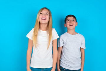 Fototapeta premium Surprised two kids boy and girl standing over blue studio background, shrugs shoulders, looking sideways, being happy and excited. Sudden reactions concept.