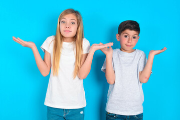 Maybe yes or no. Doubtful two kids boy and girl standing over blue studio background shrugs shoulders in bewilderment, tries to make decision puzzled what he wants. Hesitation and uncertainty.