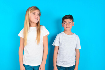 two kids boy and girl standing over blue studio background with thoughtful expression, looks away...