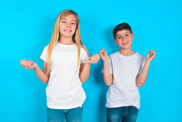two kids boy and girl standing over blue studio background, making money gesture.