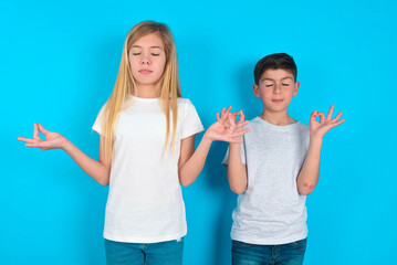 two kids boy and girl standing over blue studio background doing yoga, keeping eyes closed, holding...