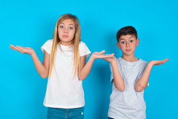 Puzzled and clueless two kids boy and girl standing over blue studio background with arms out, shrugging shoulders, saying: who cares, so what, I don't know.