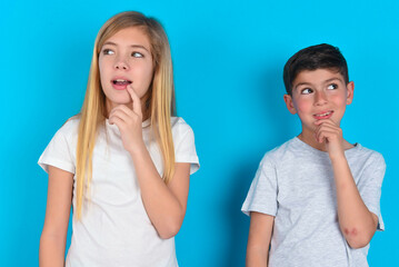 two kids boy and girl standing over blue studio background with thoughtful expression, looks to the camera, keeps hand near face, bitting a finger thinks about something pleasant.
