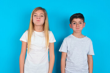 two kids boy and girl standing over blue studio background puffing cheeks with funny face. Mouth inflated with air, crazy expression.