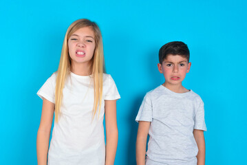 Mad crazy two kids boy and girl standing over blue studio background clenches teeth angrily, being annoyed with coming noise. Negative feeling concept.
