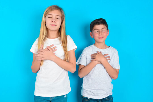 Two Kids Boy And Girl Standing Over Blue Studio Bac Closes Eyes And Keeps Hands On Chest Near Heart, Expresses Sincere Emotions, Being Kind Hearted And Honest. Body Language And Real Feelings Concept.