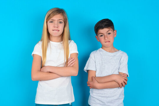 two kids boy and girl standing over blue studio background bitting his mouth and looking worried and scared crossing arms, worry and doubt.