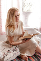 Portrait of a young beautiful fair-haired girl in a rustic style with a book near the window. A young girl is resting with a book.