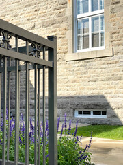 Plan rapproché d'une façade en pierre avec fenêtres d'un ancien bâtiment, clôture en fer devant des plantes à fleurs mauves. Ruelle avec un trottoir au devant une architecture antique