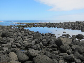 Black volcanic rocks against a deep blue sea. Small lake formed by rocks mirroring the sky. Waves, blue sky and white clouds. Rocky beach at Lehia Park, Hilo, Big Island of Hawaii.