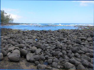 Black volcanic rocks against a deep blue sea. Waves, blue sky and white clouds. Rocky beach at Lehia Park, Hilo, Big Island of Hawaii.