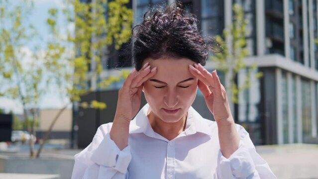 Tired exhausted young adult businesswoman, office worker having headache standing outdoors. Stressed overworked female massages head. Woman feeling terrible pain, migraine headaches, weather changes.