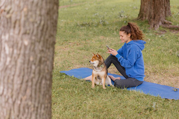a woman with a dog in the park does sports or fitness on a yoga mat. a European woman with curly hair in a blue hoodie is chatting on a smartphone and playing with a dog. a happy woman is resting