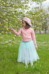 Adorable little girl stands on the green grass next to a blooming white apple tree in the park.