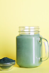 fresh green smoothie in glass bottle and spirulina powder with spoon on yellow background. useful habits, self care and healthy lifestyle. copy space