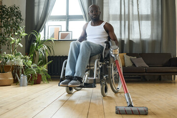 Fototapeta na wymiar African young guy sitting in wheelchair and doing housework with vacuum cleaner during rehabilitation at home