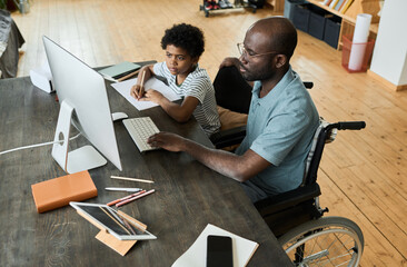 African disabled father sitting in wheelchair using computer at table to help his son with homework