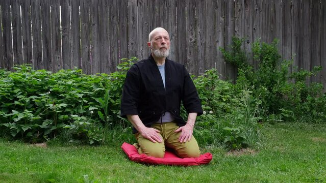 bald and bearded senior man wearing a short kimono is sitting in a traditional Japanese seiza position on a cushion pad in a backyard and practicing deep box breathing