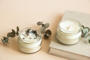 Two candmade candles from paraffin and soy wax in glass with wooden wick and dry herbal isolated on pastel beige background. Flat lay, top view, copy space - obrazy, fototapety, plakaty
