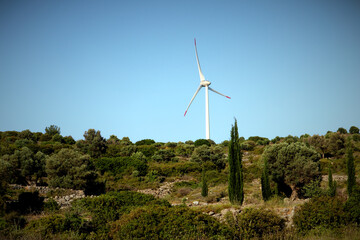 A wind turbine (windmill) is standing lonely on a hilltop in the middle of Aegean vegetation. In...