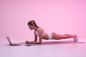 Young woman in neon light standing in plank position, watching online workout tutorial on laptop