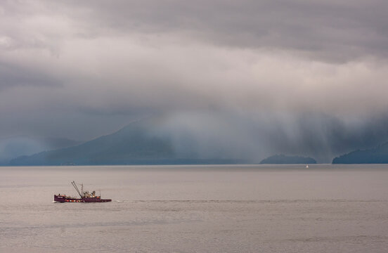 Hoonah, Alaska, USA - July 18, 2011: Deep lying marroon Ocean Maid fishing vessel moving in brown water while foggy clouds turn into rain over partly visible forested mountains on horizon.