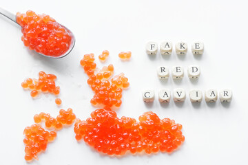 Fake red caviar eggs on a white surface next to a spoon and the inscription fake red caviar. Concept of cheating the consumer and selling counterfeit food products. - obrazy, fototapety, plakaty