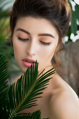 Beautiful young mixed-race woman with professional makeup and perfect skin posing with tropical green leaves. SPA and wellness, body care and skincare.  Close up, selective focus