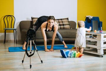 Young mother fitness instructor personal trainer record video near camera at home with baby daughter