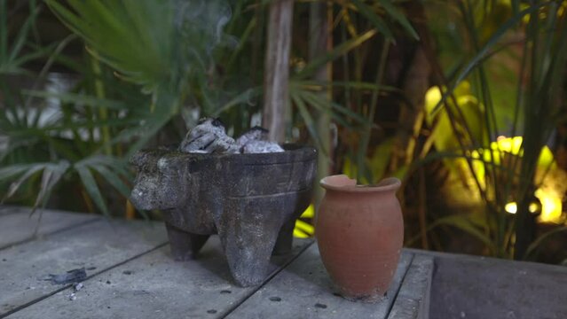 Smoking Incense burner with coal and pottery in tropical garden  with palm trees and yellow light in the background in Tulum