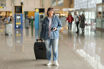 smiling middle aged woman with suitcase and passport standing in arrival or departure hall of an...