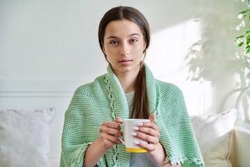 Young teenage female under knitted plaid warming up with hot tea in mug, looking at camera