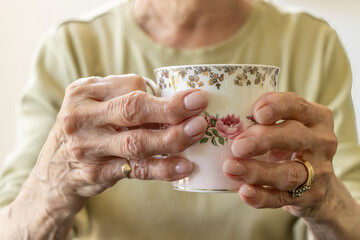 Image of an elderly woman with arthritis in her hands trying to hold a beautiful tea cup. Her fingers are twisted and contrast against the delicate tea cup. - obrazy, fototapety, plakaty