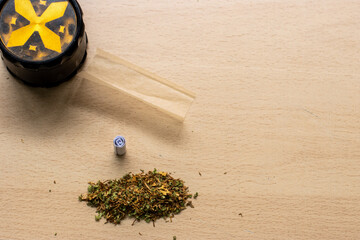 Preparing a joint and drug paraphernalia concept theme with herb girder used to grind cannabis buds and roll marijuana joints, next to rolling paper and weed bud isolated on wooden background - obrazy, fototapety, plakaty