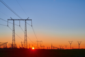 Orange sunrise on field with high voltage electricity towers. Dark silhouettes of repeating power lines on sunrise. Electricity generation, transmission, and distribution network. Industrial landscape